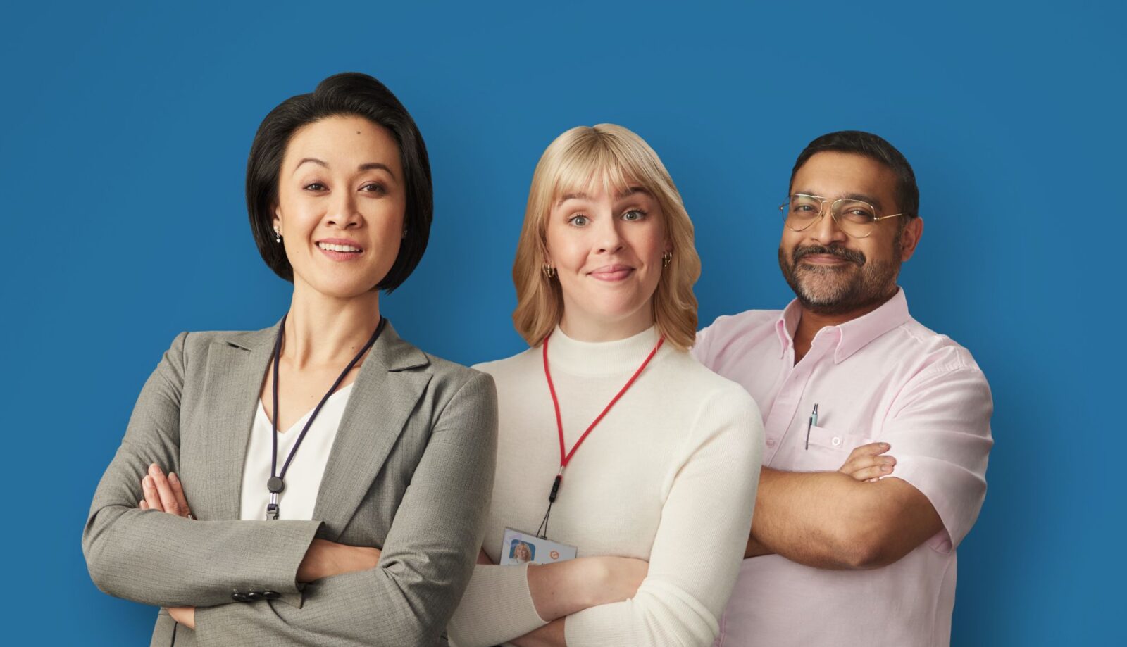 Three professionals stand confidently in front of a blue background. The woman on the left wears a grey suit, the woman in the middle wears a white top, and the man on the right wears glasses and a light pink shirt.