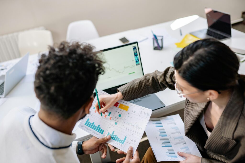 Two people review printed charts and graphs at a desk with an open laptop displaying data. Office supplies are scattered on the white table around them. Both are focused on the documents.
